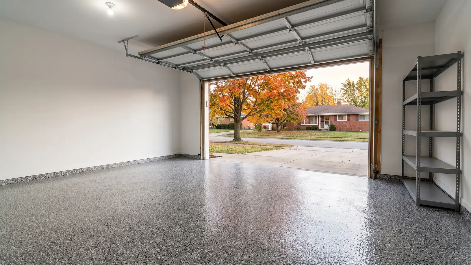 Empty two-car garage with a glossy gray, black and white flake epoxy floor, the overhead door open to a driveway and an autumn maple