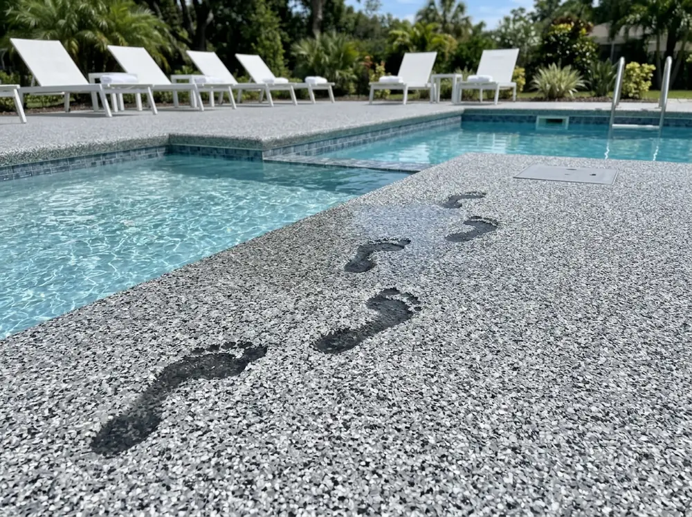 Pool deck coated in a gray and white flake system with a fine grit texture, wet footprints near the pool edge, lounge chairs in the background